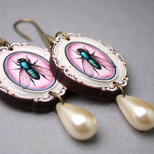 Puede incluir: Un par de pendientes con un marco ovalado rosa y blanco con un insecto azul y verde. Cada pendiente tiene una gota de perla blanca.