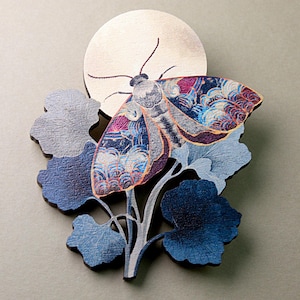 May include: A decorative wooden moth with intricate wing patterns in blue, purple, and orange, set against a silver moon and blue foliage. The artwork is mounted on a grey background.
