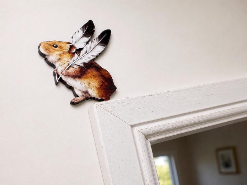 May include: A decorative wall cutout featuring a brown and tan hamster with large, white, black-tipped feathers attached to its back. The hamster is depicted in profile, facing left, with its front paws holding the base of the large white feathers.