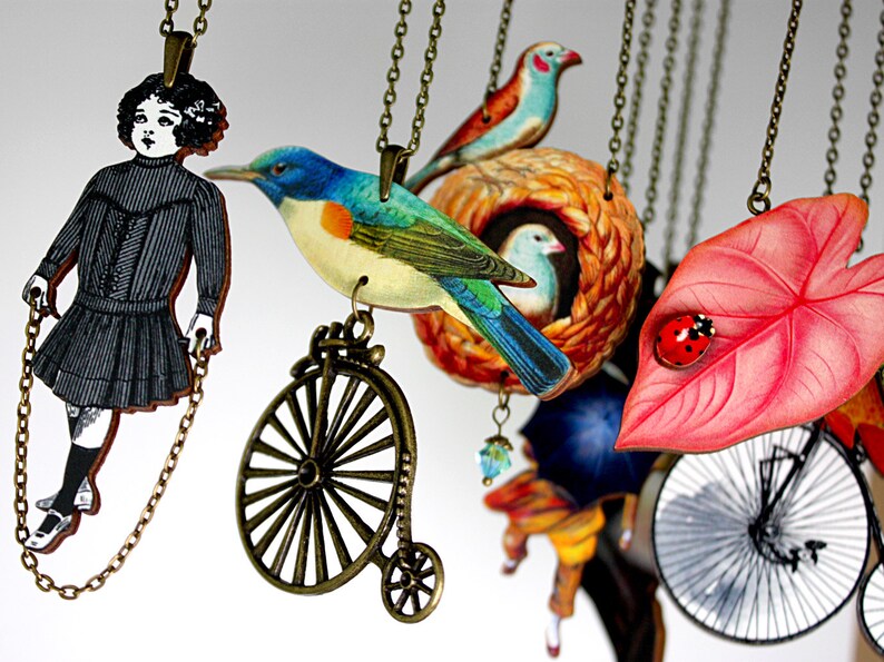 May include: A collection of necklaces with various charms, including a girl in a black dress, a blue bird, a bird's nest, a leaf with a ladybug, and a bicycle. The necklaces are made of metal chains and have a vintage aesthetic.