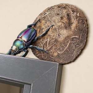 Puede incluir: Un adorno decorativo de pared que presenta un escarabajo tridimensional iridiscente con un caparazón metálico morado y verde, colocado junto a un disco marrón circular texturizado que parece una bola de materia orgánica seca.