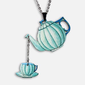 Könnte beinhalten: Eine Halskette mit einer teeblauen Teekanne und einer Teetasse. Die Teekanne hat eine silberne Kette, die am Ausguss befestigt ist und mit der Teetasse verbunden ist. Die Teetasse steht auf einem Unterteller.