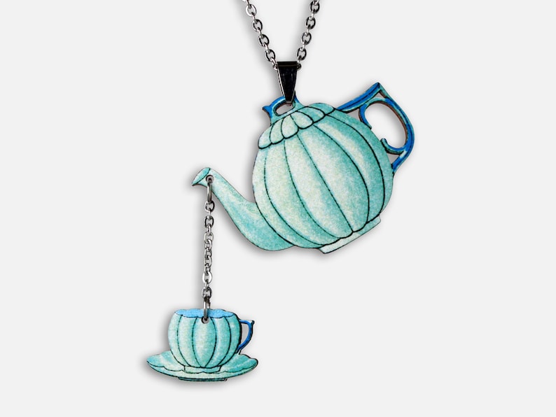May include: A teal blue teapot and teacup necklace. The teapot has a silver chain attached to the spout, which connects to the teacup. The teacup is on a saucer.
