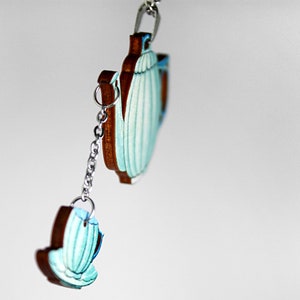 May include: A teal blue teapot and teacup charm with a silver chain. The teapot and teacup are made of wood and have a painted design.