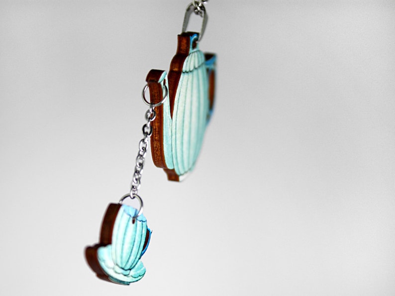 May include: A teal blue teapot and teacup charm with a silver chain. The teapot and teacup are made of wood and have a painted design.