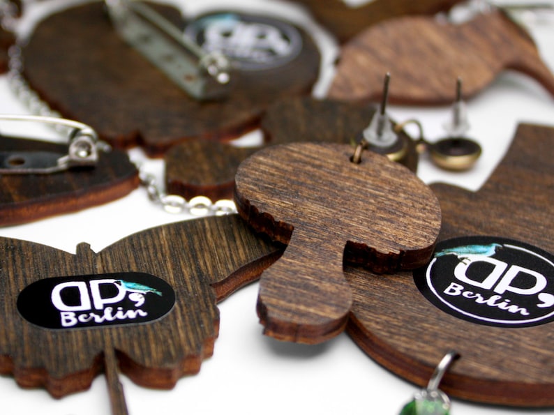 May include: A set of wooden earrings featuring a mushroom, butterfly, and bird designs. The earrings are brown and have a natural wood grain finish. Each earring has a small metal pin and a black sticker with the text "dp, Berlin".