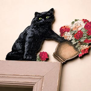 Puede incluir: Una decoración de pared de un gato negro con ojos de color verde brillante, colocado como si estuviera volcando un jarrón lleno de un ramo vibrante de flores rojas, rosas y blancas. El gato y el jarrón son piezas separadas sobre un marco.