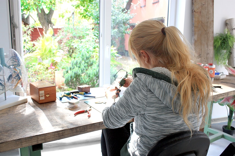 May include: A person with blonde hair tied in a ponytail wears a grey and white heathered zip-up hoodie while working at a wooden desk. The desk holds a small green handheld rotary tool, red-handled pliers, a wooden box, and various craft supplies.