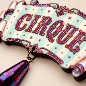 May include: A decorative sign featuring the word CIRQUE in purple lettering on a light blue and cream striped background with small pink stars. The sign has an ornate purple border and a single, iridescent, teardrop-shaped pendant hanging from it.