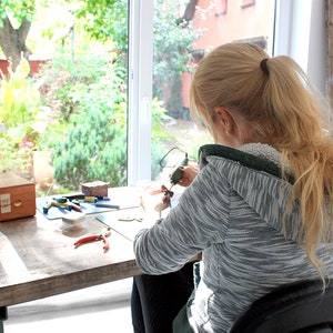 May include: A woman sits at a table working on a craft project. She is using a small tool to work on a piece of material. There are various tools and supplies on the table, including a wooden box, pliers, and a blue cutting mat.