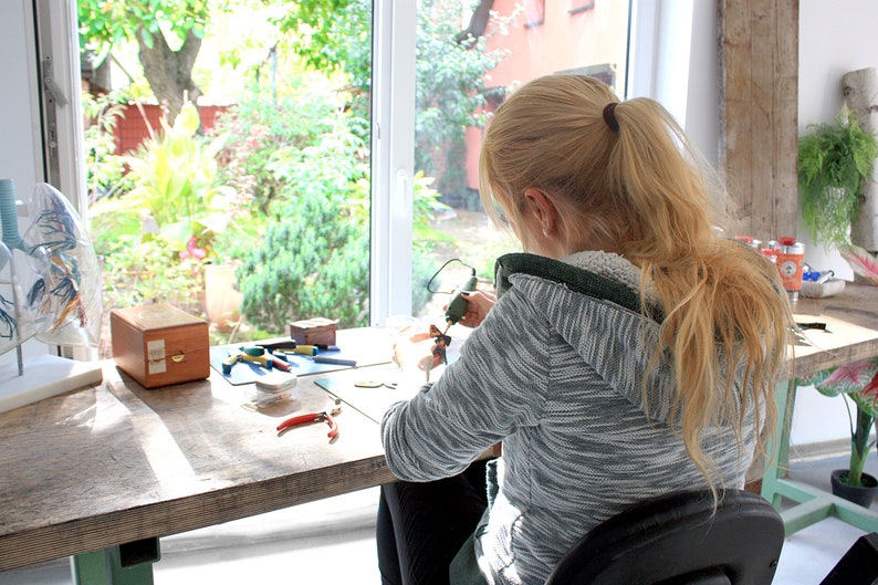 May include: A woman sits at a table working on a craft project. She is using a small tool to work on a piece of material. There are various tools and supplies on the table, including a wooden box, pliers, and a blue cutting mat.
