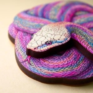 Könnte beinhalten: Eine farbenfrohe Schlangenskulptur mit detailliertem Design. Die Schlange ist gewunden und weist Farbtöne von Lila, Blau und Rosa auf. Der Kopf ist weiß mit blauen Akzenten und lila Augen. Die Zunge ist rosa, und die Ränder sind dunkelbraun.