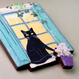 Puede incluir: Un gato negro se sienta en un alféizar de ventana mirando por una ventana con un marco azul. La ventana está pintada con un fondo amarillo y tiene un marco blanco. Una pequeña maceta morada con flores blancas está a la derecha del gato.