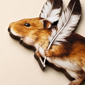 May include: A flat wooden cutout shaped like a hamster with brown and white fur. The hamster is depicted holding two large, detailed white feathers with dark brown tips that extend upwards from its body, creating a wing-like appearance.