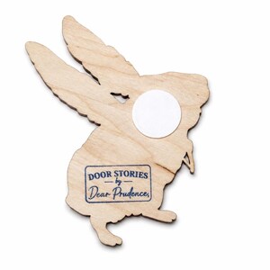 May include: A light-coloured wooden cutout shaped like a rabbit with long ears. A circular white adhesive patch is attached to the upper body. A rectangular blue ink stamp on the lower body reads DOOR STORIES by Dear Prudence.