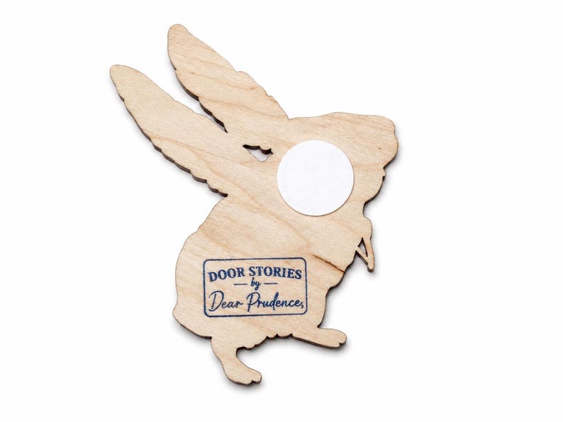 May include: A light-coloured wooden cutout shaped like a rabbit with long ears. A circular white adhesive patch is attached to the upper body. A rectangular blue ink stamp on the lower body reads DOOR STORIES by Dear Prudence.