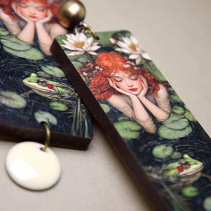 Op de afbeelding: Een paar rechthoekige houten oorbellen. Elke oorbel heeft een kleurrijk beeld van een vrouw met rood haar, liggend in een vijver met waterlelies en kikkers. De oorbellen hebben een witte keramische hanger die aan de onderkant is bevestigd.