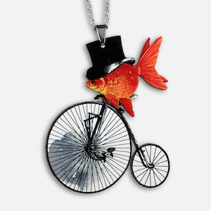Könnte beinhalten: Ein Anhänger in Form eines goldenen Fisches, der einen Zylinder trägt und ein Hochrad fährt. Der Fisch ist orange und rot mit schwarzen Augen. Das Fahrrad ist schwarz mit weißen Reifen. Die Halskette ist silber.