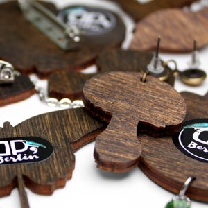 May include: A set of wooden earrings featuring a mushroom, a butterfly, and a bird design. The earrings are brown and have a natural wood finish. The butterfly earring has a black and white logo that says "dp, Berlin".