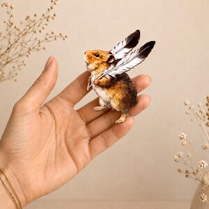 May include: A small, detailed figurine of a brown and white hamster with two large, black and white feathers attached to its back, resembling wings. The hamster is depicted in a standing position with its mouth slightly open, showing fine fur texture.