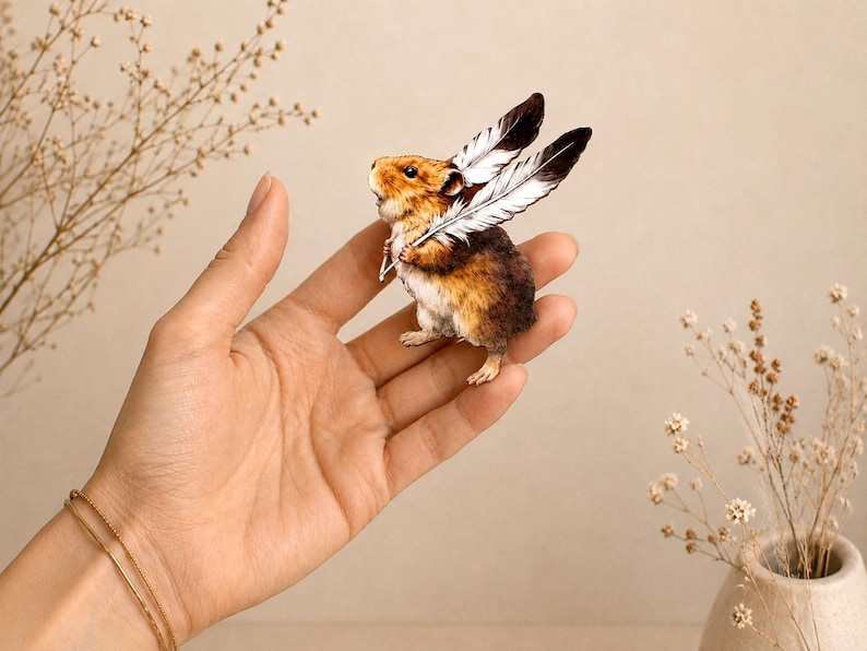 May include: A small, detailed figurine of a brown and white hamster with two large, black and white feathers attached to its back, resembling wings. The hamster is depicted in a standing position with its mouth slightly open, showing fine fur texture.