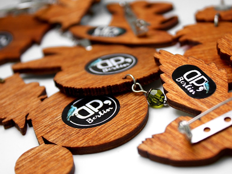 May include: A collection of wooden pins with a circular design featuring the text "DP, Berlin". The pins are in various shapes and sizes, and some have a silver pin back.