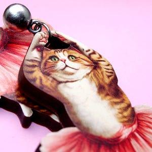 May include: A whimsical cat-themed earring featuring a tabby cat in a ballet pose. The cat has green eyes and is wearing a pink tutu. The earring is attached to a silver-colored ball and a metal hoop.