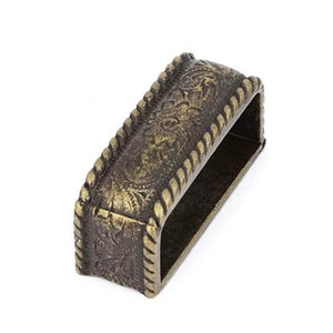 Può includere: Una fibbia in metallo dorato con un bordo a forma di corda e un intricato disegno floreale. La fibbia è rettangolare e ha una finitura liscia e lucida.