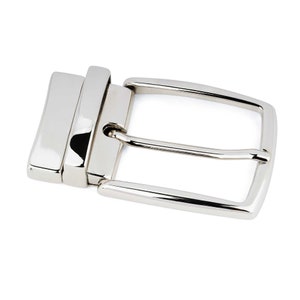 Puede incluir: Una hebilla de cinturón plateada con un acabado liso y pulido. La hebilla tiene una forma rectangular con un borde redondeado y una sola lengüeta.