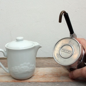 May include: A white ceramic creamer with a lid and a silver and black stovetop espresso maker with a black handle and a copper spout.