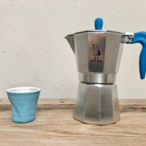 May include: A silver coffee maker with a blue handle and lid. The coffee maker has a logo that says "ATTIRUM" and a small illustration of a person. A light blue ceramic cup is in the foreground.