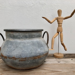 May include: A large, grey metal planter with two handles. The planter is sitting on a wooden surface. A wooden artist's mannequin is standing behind the planter.