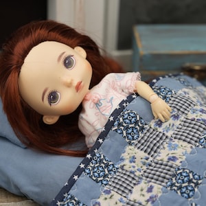 Peut inclure: Une poupée aux cheveux roux et aux yeux bleus est allongée dans une courtepointe patchwork bleue et blanche. La poupée porte une robe rose et blanche.