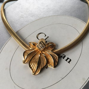 May include: Gold-toned necklace featuring a floral pendant with a central square of small, clear crystals. The necklace has a mesh-like chain. The pendant is a stylized flower with detailed petal design.