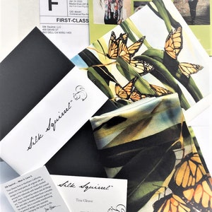 May include: A close-up of a silk scarf featuring a colourful butterfly design. The scarf is folded and displayed on a white surface with a business card that reads "Silk Squirrel" and "Tina Gleave".