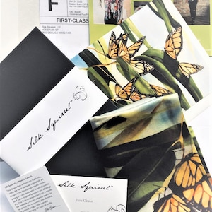 May include: A close-up of a silk scarf with a colorful butterfly design. The scarf is folded and displayed on a white surface with a business card that reads "Silk Squirrel" and "Tina Gleave".