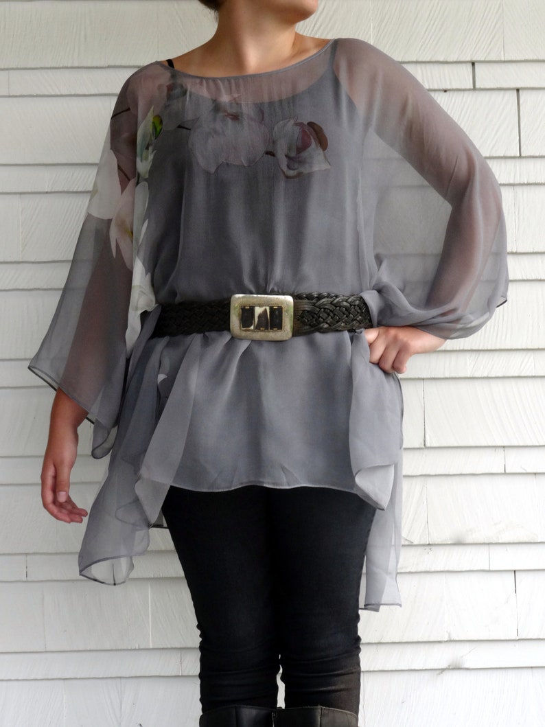 May include: A gray sheer top with a floral print and a brown woven belt. The top has long sleeves and a loose fit. The model is wearing black pants.