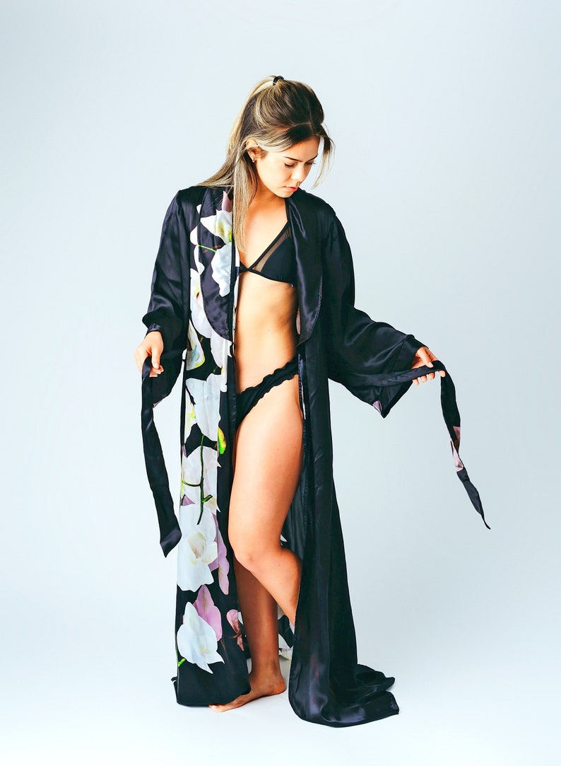 May include: A black satin robe with a floral print. The robe has long sleeves and a tie belt. The model is wearing a black bra and underwear.