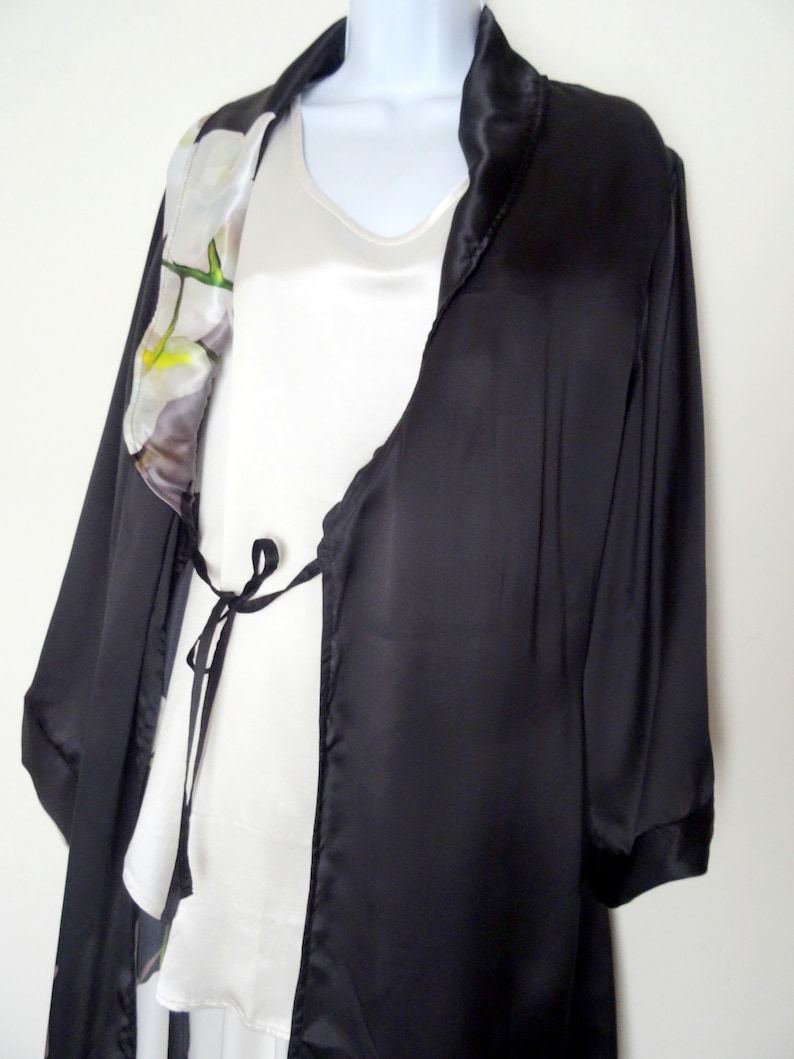 May include: A black satin robe with a white floral print on the collar. The robe has a tie at the waist and long sleeves.