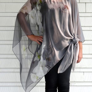 May include: A gray sheer top with white floral print. The top has a loose, flowing fit and is tied at the waist. The model is wearing black pants and boots.