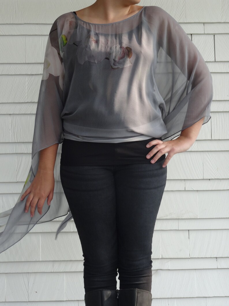 May include: A gray sheer top with a floral print and a long, flowing design. The top is worn with black jeans.