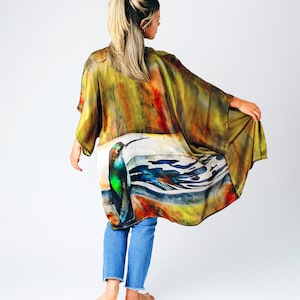 Pu&ograve; includere: Un lungo kimono di seta fluida con un design ad acquerello di un colibr&igrave; e forme astratte in tonalit&agrave; di verde, blu, arancione e giallo. Il kimono &egrave; indossato sopra un paio di jeans blu.