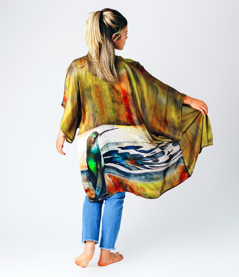 Pu&ograve; includere: Un lungo kimono di seta fluida con un design ad acquerello di un colibr&igrave; e forme astratte in tonalit&agrave; di verde, blu, arancione e giallo. Il kimono &egrave; indossato sopra un paio di jeans blu.