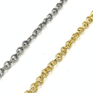 May include: Two chains, one silver and one gold, are laid out on a white surface. The chains are both made of small, linked circles.