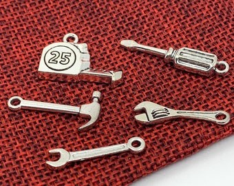 May include: A set of five silver-toned metal charms shaped like tools. The charms include a tape measure with the number 25 on it, a hammer, a wrench, a screwdriver, and a pair of pliers.