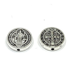 Peut inclure: Deux pendentifs ronds de couleur argentée. L'un présente une figure avec du texte sur le bord. L'autre a une croix et des lettres. Les deux ont un petit trou pour l'enfilage.