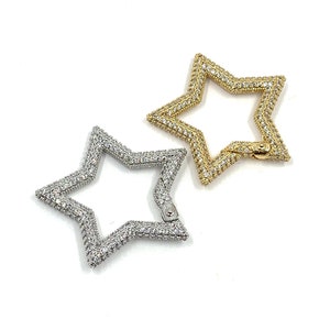 May include: Two star-shaped clasps, one gold-toned and one silver-toned, are displayed against a white background. Both clasps are encrusted with small, clear gemstones, creating a sparkling effect. The gold clasp is open, while the silver clasp is closed.