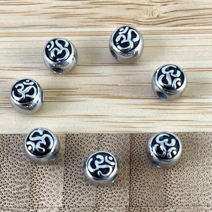 May include: Seven silver-colored beads with a black and white Om symbol design. The beads are round and have a hole for stringing. They are arranged on a light brown bamboo surface.