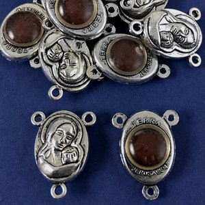 May include: A group of silver-toned metal charms with a three-dimensional image of the Virgin Mary holding a baby Jesus. Each charm has a round, brown glass insert with the words "TERRA" and "JERUSALEM" printed on it.