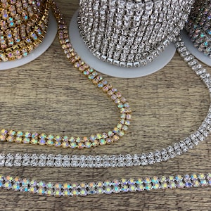 K&ouml;nnte beinhalten: Drei Spulen mit Strassbesatz, eine gold, eine silber und eine regenbogenfarben. Der Besatz ist zweireihig und liegt auf einer Holzoberfl&auml;che.